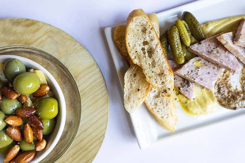 dish of olives and almonds next to a plate of bread, cornichons, and pate
